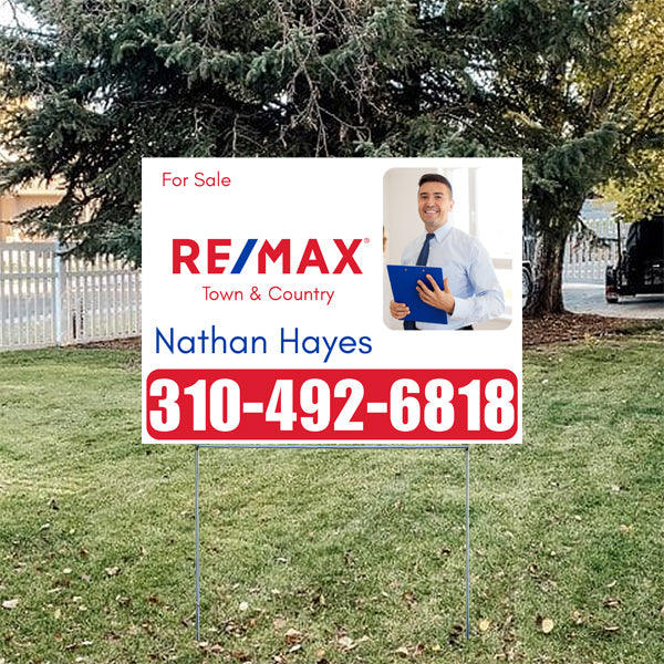 Real estate yard sign for RE/MAX Town & Country featuring a smiling real estate agent in a white shirt holding a clipboard, with "For Sale" text, contact name Nathan Hayes, and phone number 310-492-6818, set against a grassy lawn with trees in the background.
