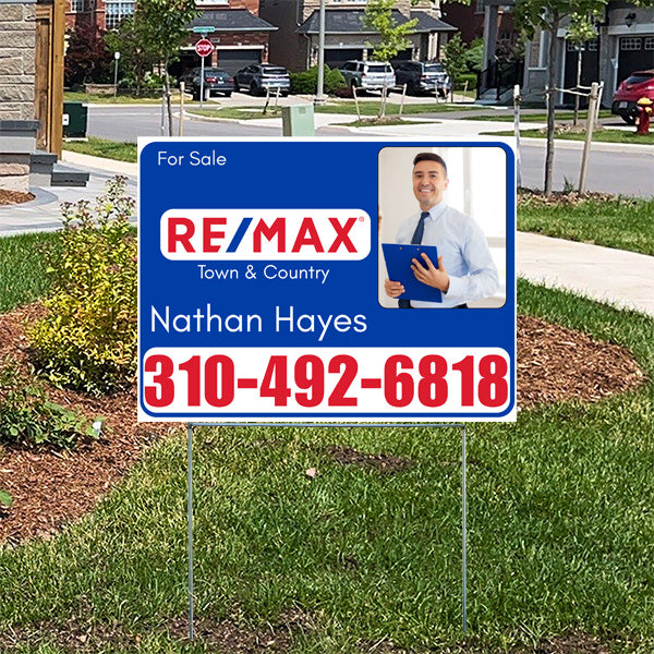 Real estate for sale sign featuring RE/MAX Town & Country, contact agent Nathan Hayes at 310-492-6818, with an image of a smiling man holding a folder, placed on a residential neighborhood lawn.