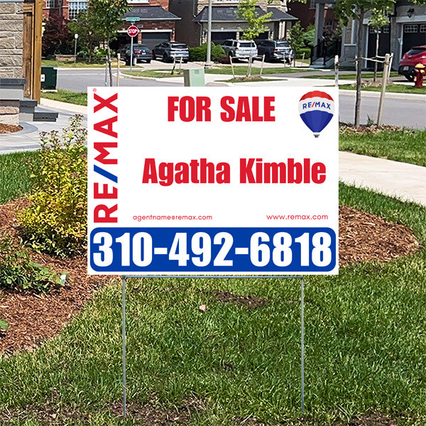 Real estate for sale sign on a lawn by RE/MAX with agent name Agatha Kimble and contact number 310-492-6818, set in a suburban neighborhood.