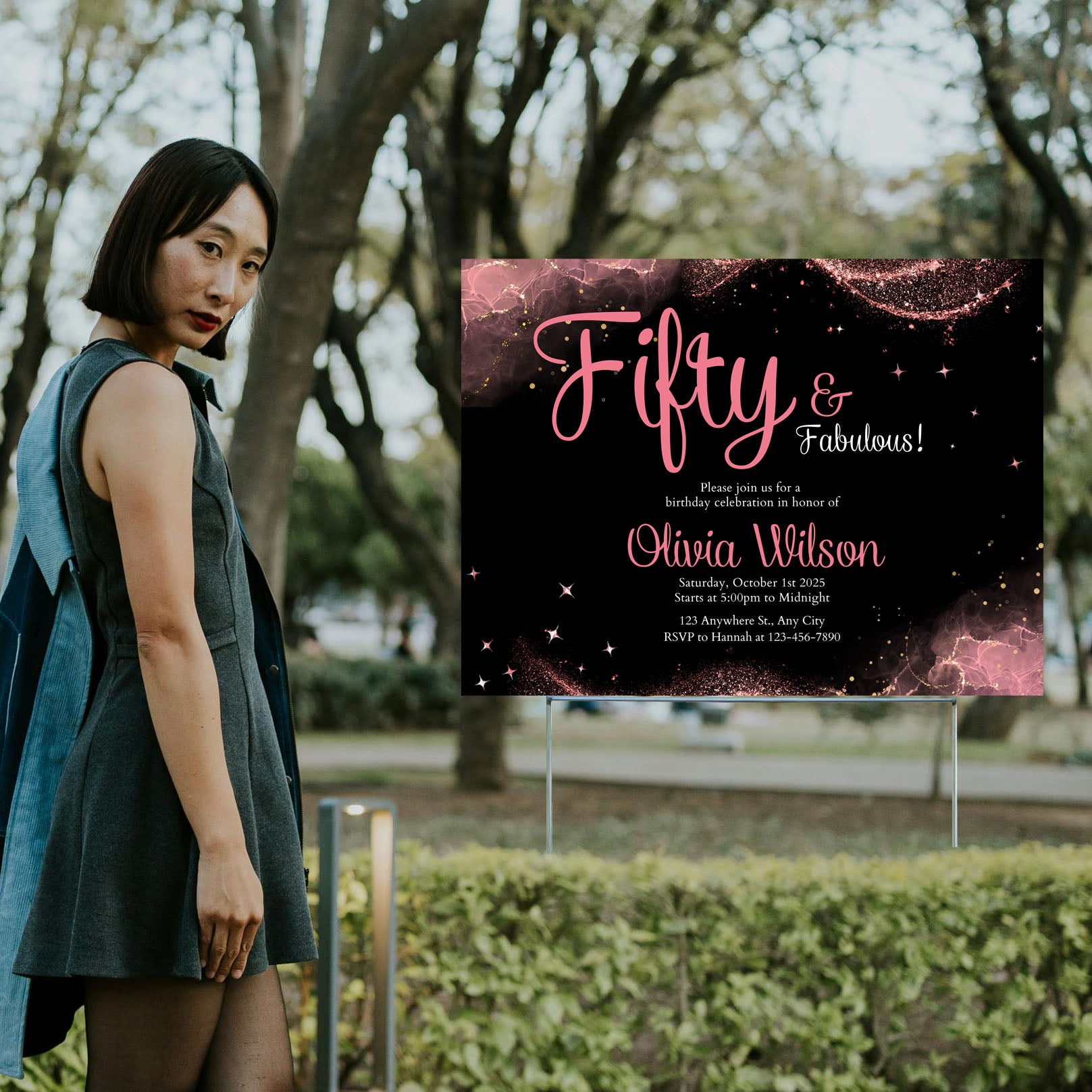 Woman in a gray dress standing in a park next to a birthday invitation sign reading "Fifty & Fabulous" with event details.