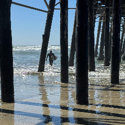 Person walking along a beach under a pier with waves in the background