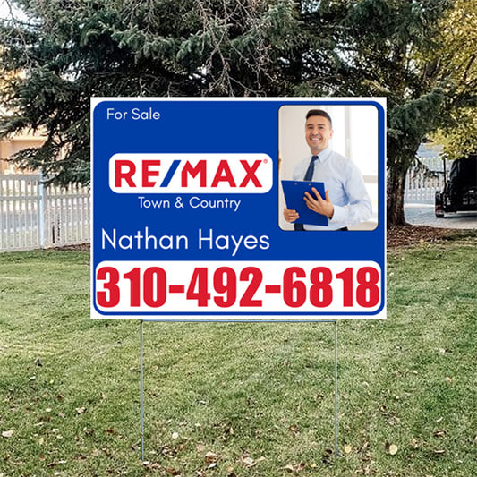 Real estate for sale sign with RE/MAX logo, contact number, and real estate agent photo, located on a grassy lawn with trees in the background.