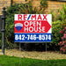 Real estate open house sign for RE/MAX with contact number 842-746-8574 placed in a landscaped yard.